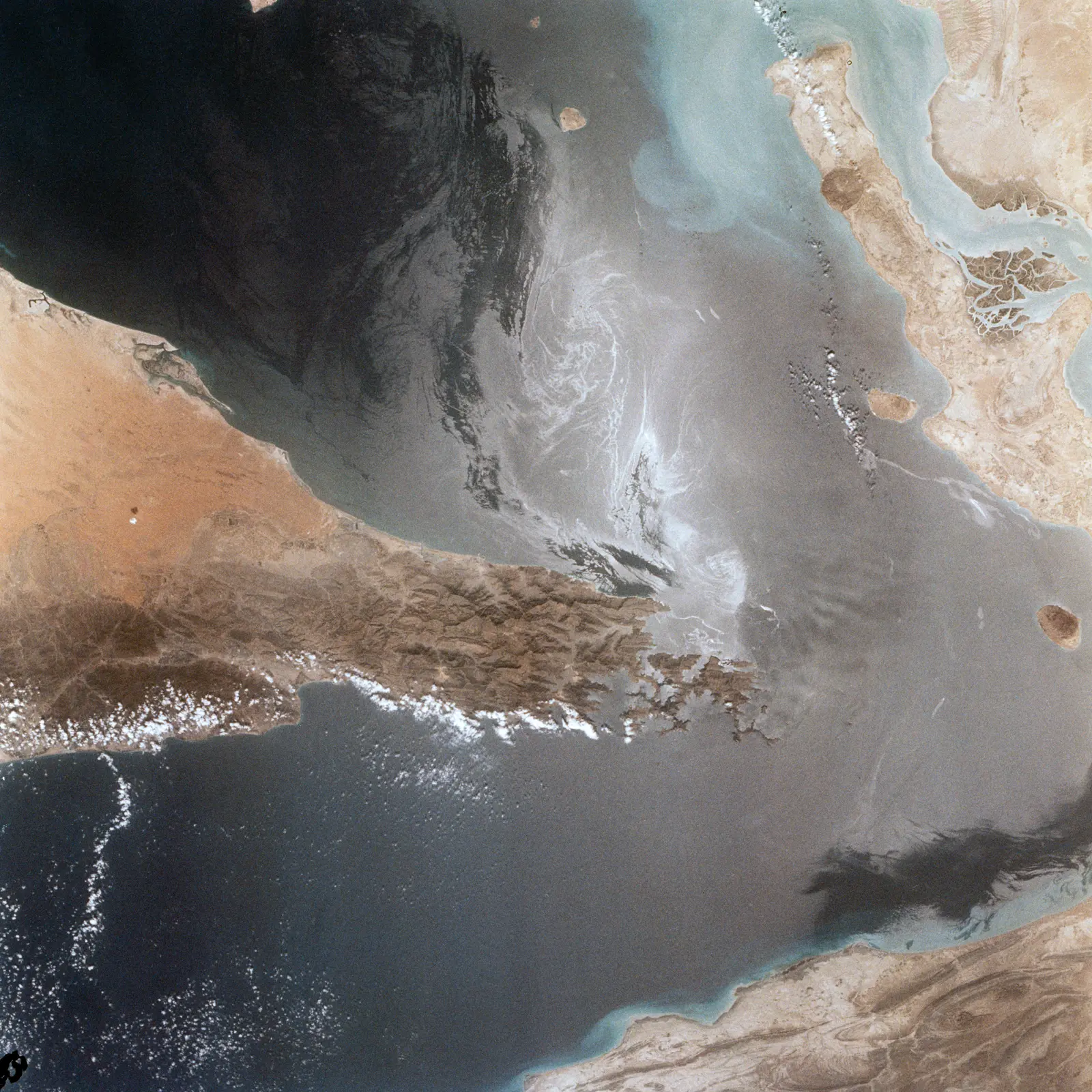 A satellite image shows a narrow body of water bordered by arid, brown landmasses, with swirling white patterns visible on the dark water's surface. The photographer titled it “Strait of Hormuz”.