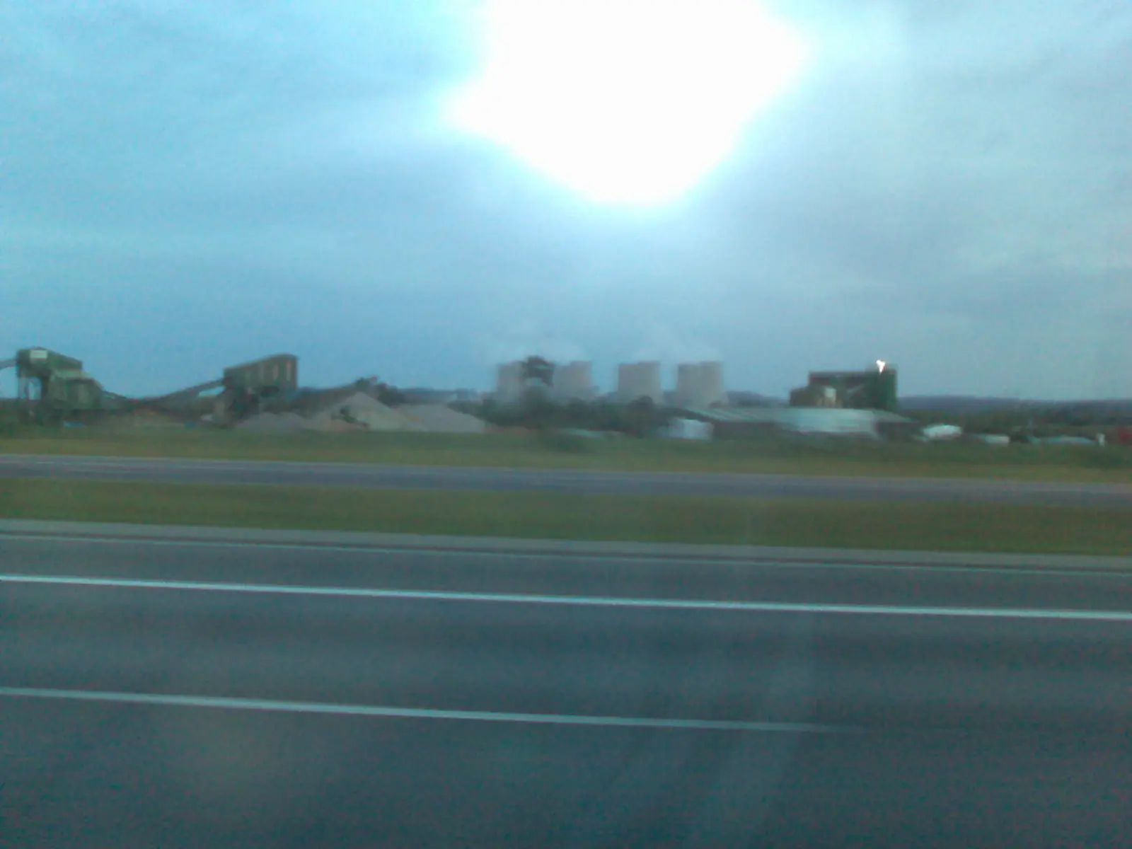 A view from a highway shows power plant cooling towers and industrial buildings in the distance under a cloudy sky. The photographer titled it “Big power plant cooling towers”.