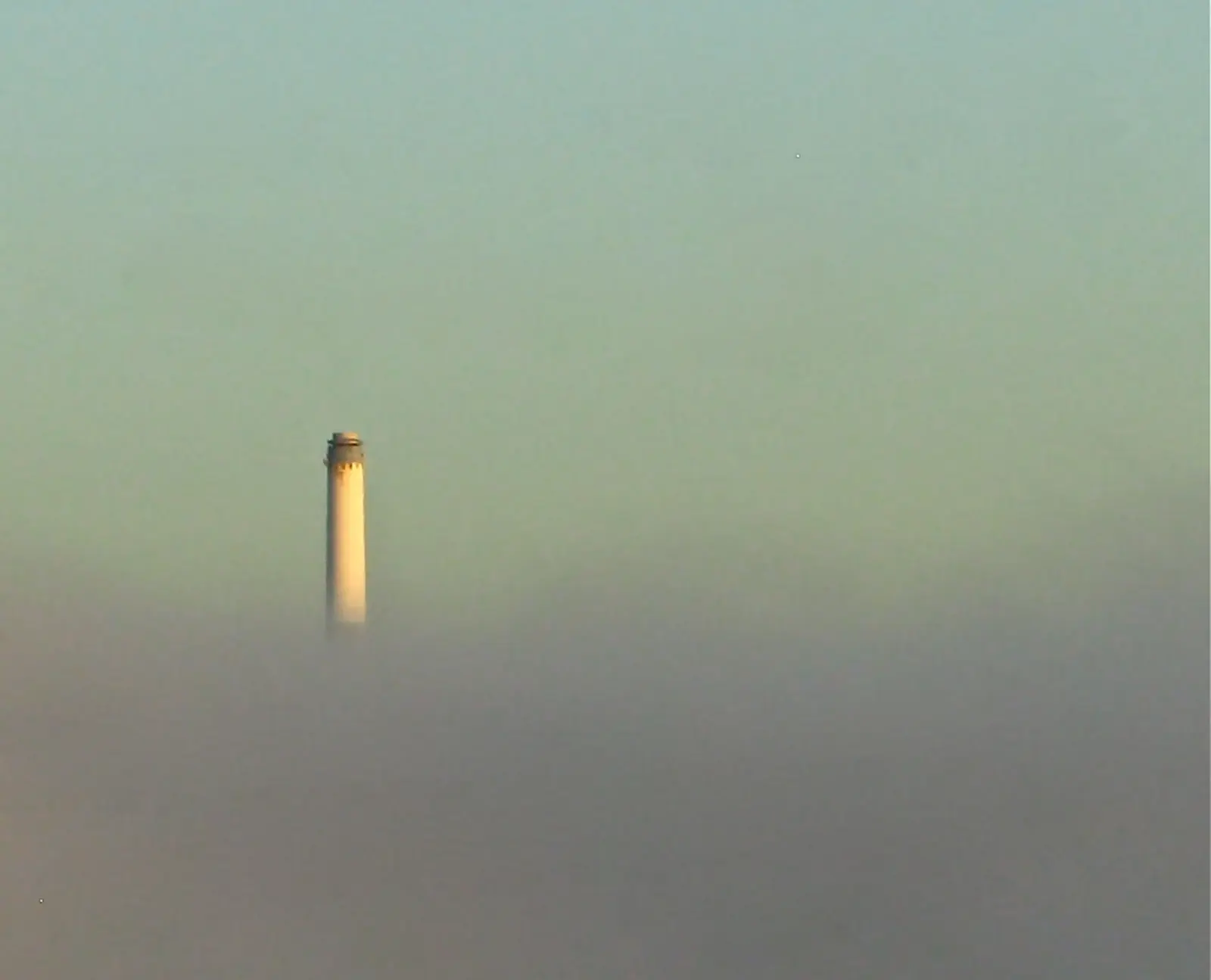 A white lighthouse emerges from thick fog beneath a pale sky. The photographer titled it “Morning Drive”.