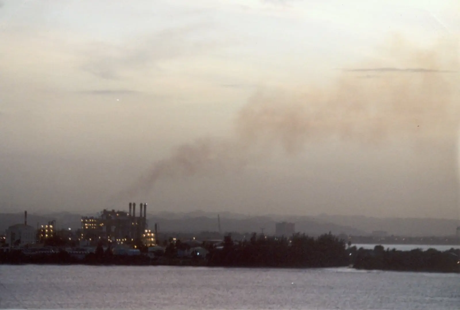 A factory complex with multiple smokestacks emitting a plume of smoke sits on the edge of a body of water under a dim sky.