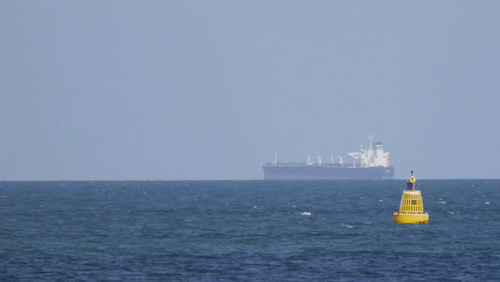A distant cargo ship sails on the horizon of a blue sea, with a yellow buoy floating in the right foreground. The photographer titled it “Distant Cargo Vessel”.