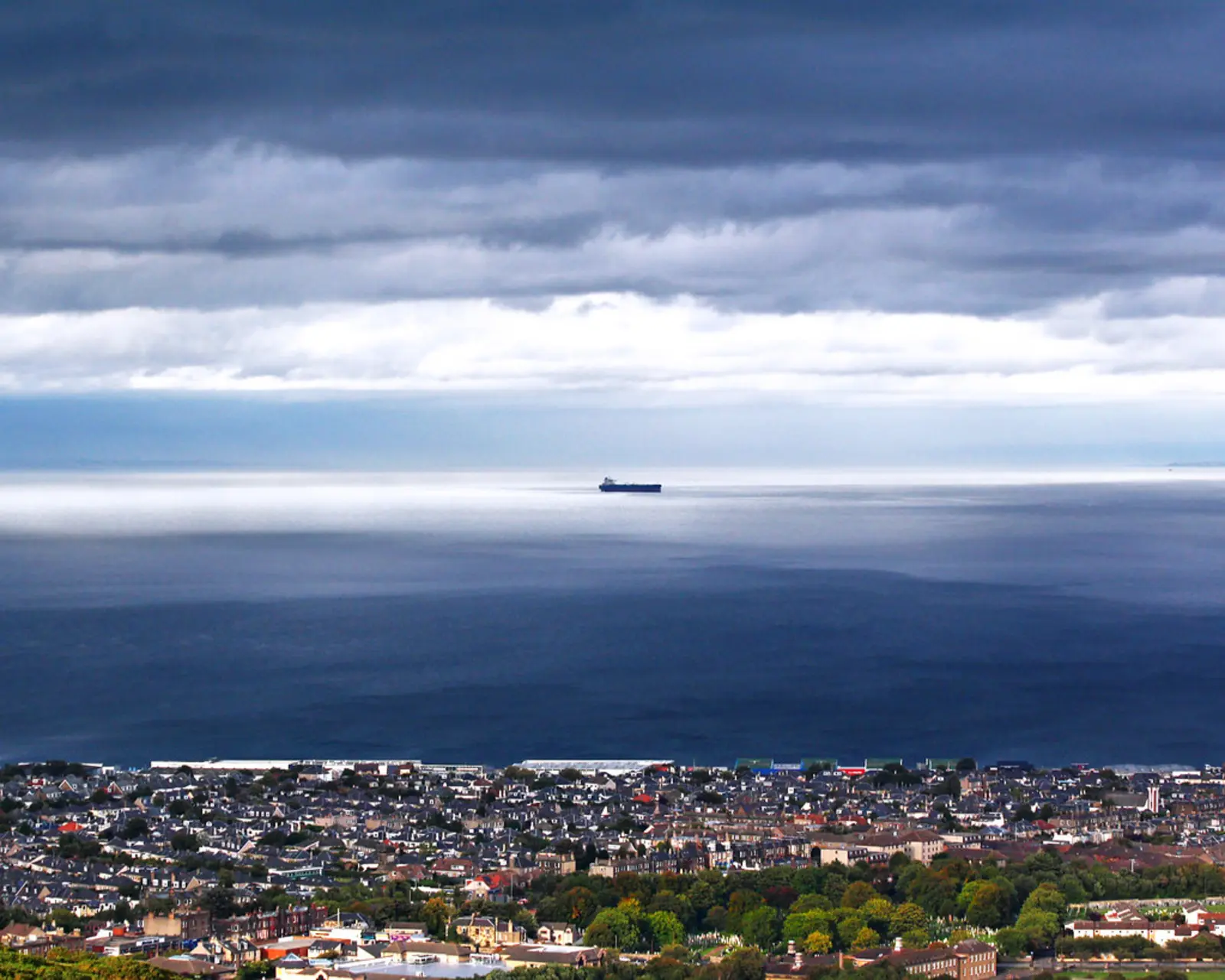 A sprawling coastal town sits in the foreground beneath a vast, dark body of water, where a single ship is visible on the horizon under a heavily clouded sky. The photographer titled it “Firth of Forth at sunset, Edinburgh, Scotland”.