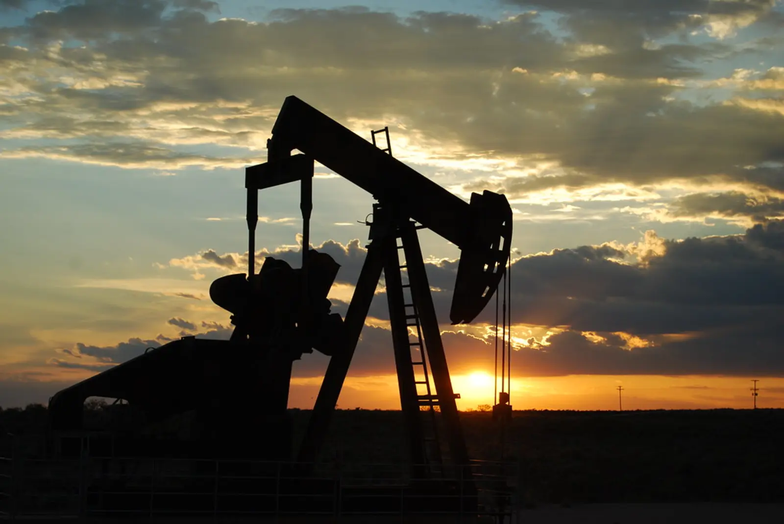 A silhouetted oil pump jack stands in a flat landscape against a cloudy sunset sky.