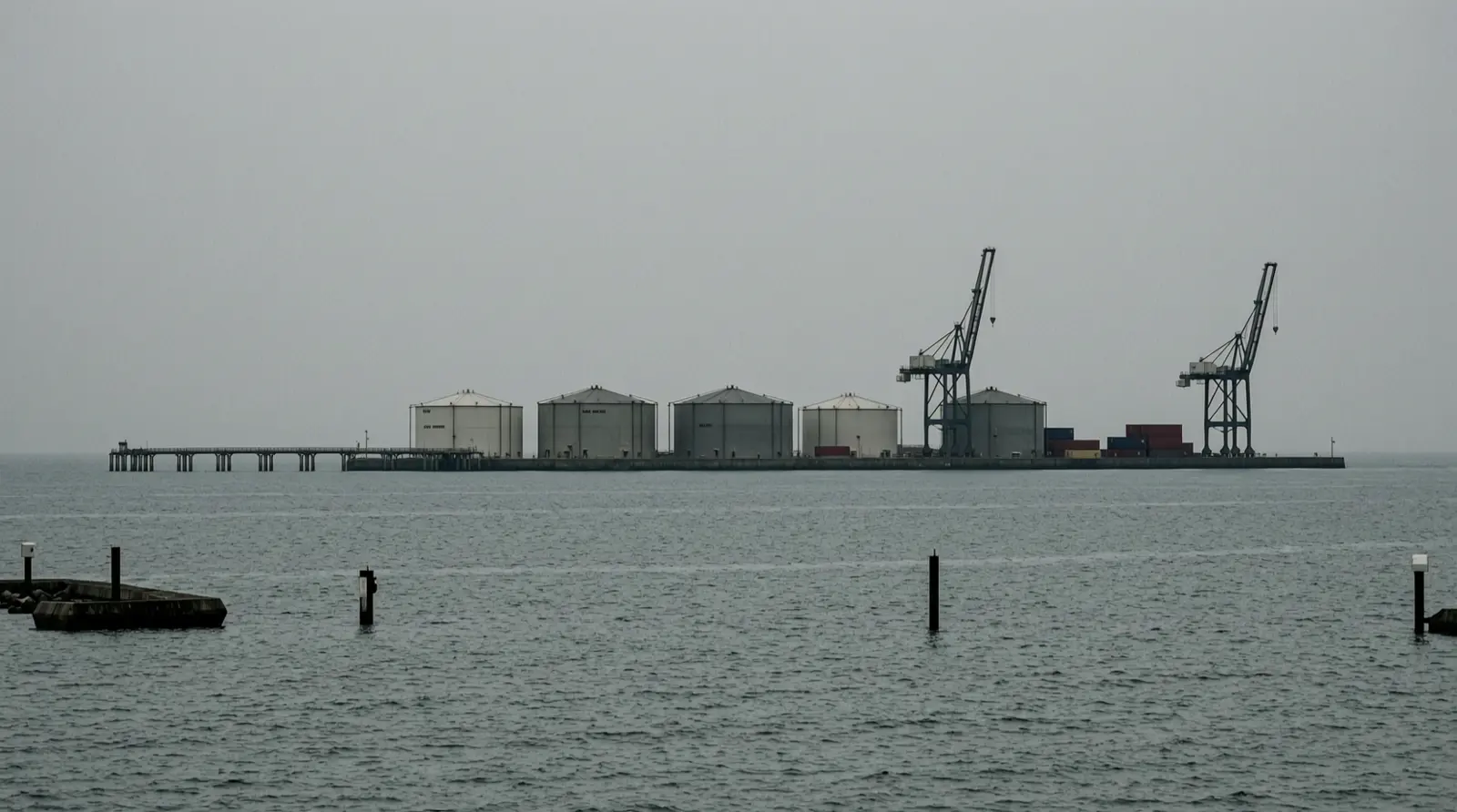 A coastal oil storage terminal seen across water, with tanks and a jetty in haze.