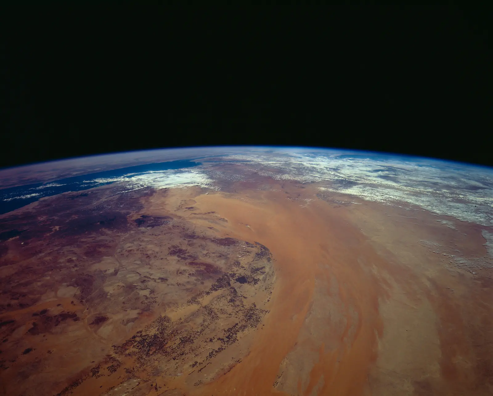A view from space shows the curvature of the Earth above a vast expanse of brown and tan desert terrain with scattered white clouds. The photographer titled it “Northern Saudi Arabia as seen from STS-62”.
