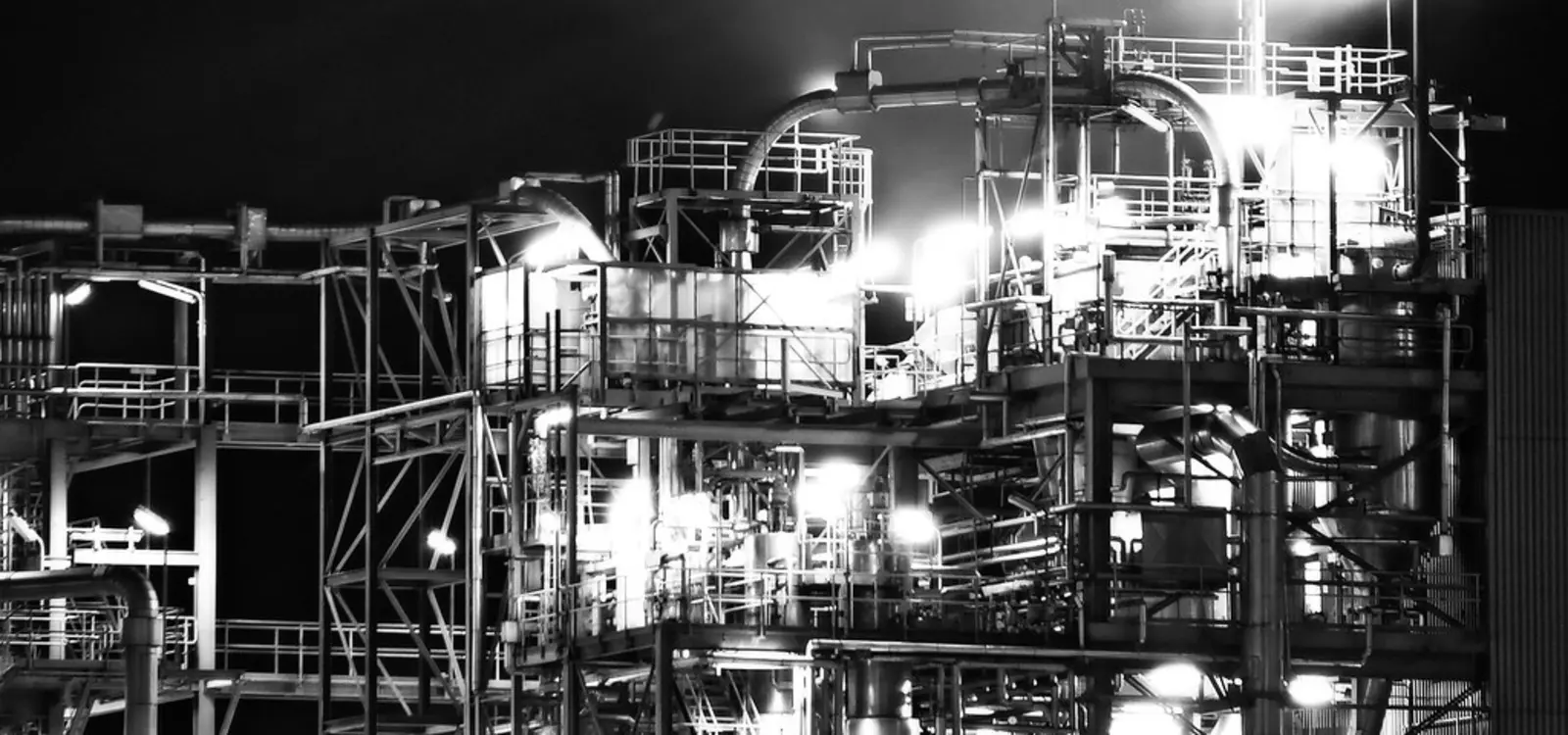 A black-and-white photograph of a complex industrial facility at night, illuminated by bright lights and featuring extensive networks of pipes and metal structures. The photographer titled it “POPS AND PIPES 2”.
