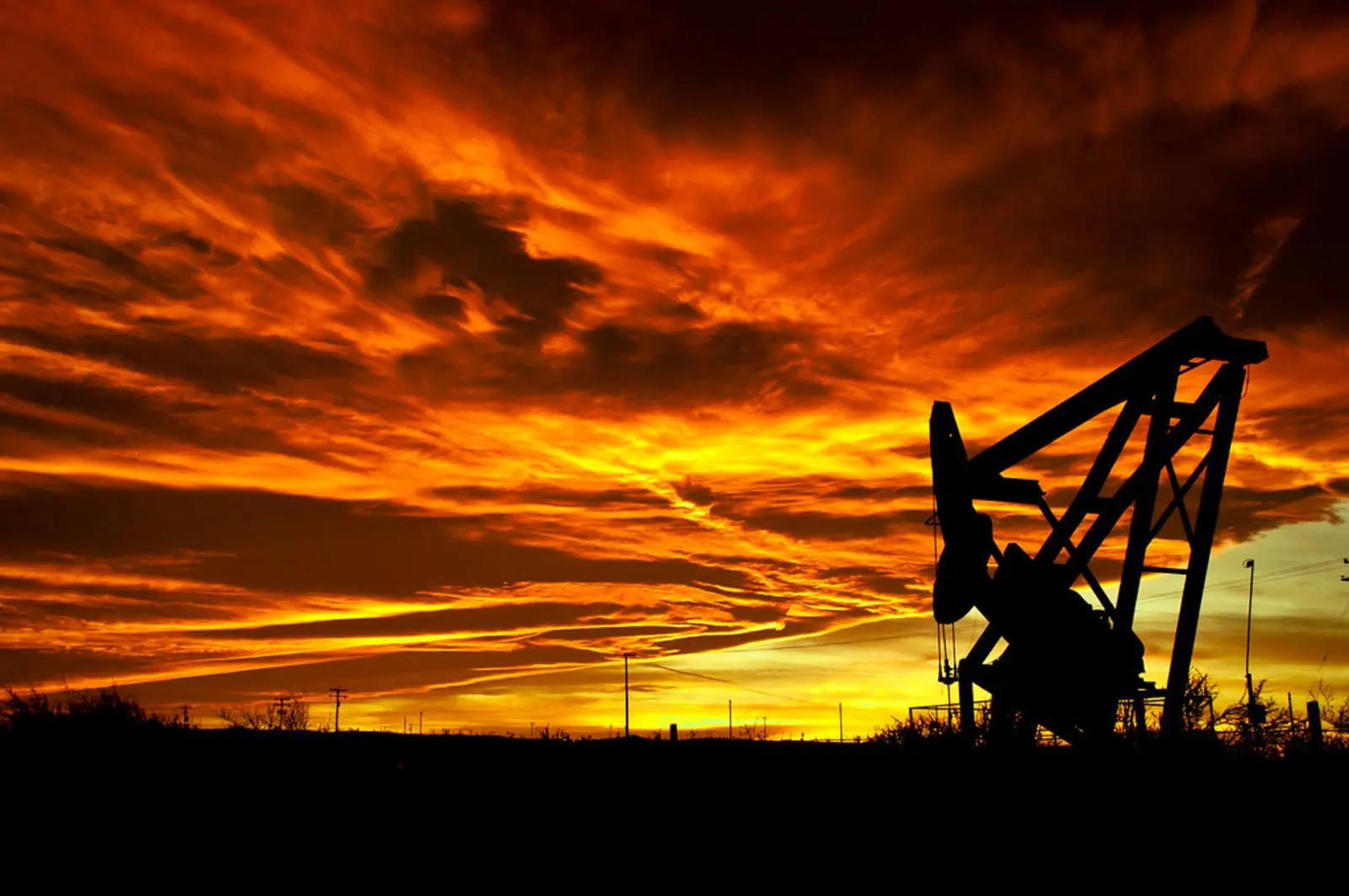 A silhouetted oil pump jack stands in a field against a dramatic, fiery orange sky. The photographer titled it “The Burning of the Sky”.