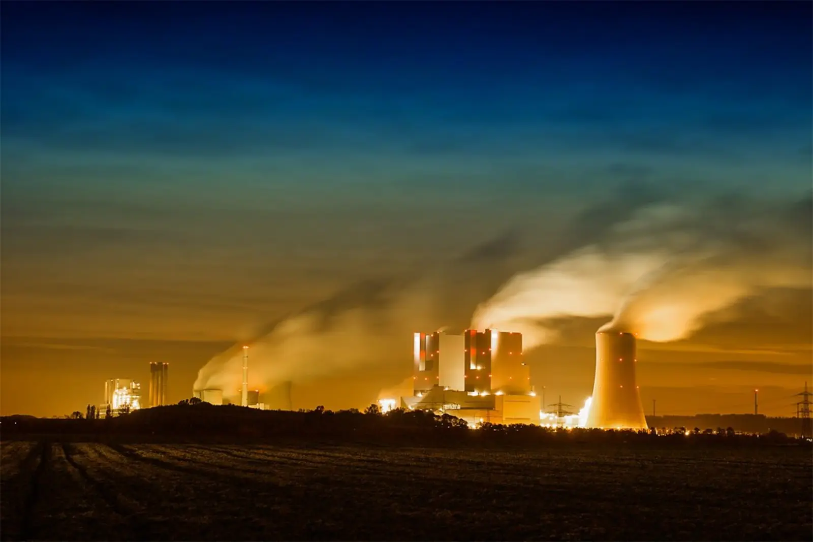 A large power plant with cooling towers emits thick plumes of steam into the dark, twilight sky, illuminated by orange facility lights. The photographer titled it “Air pollution - Credit to http://homedust.com/”.