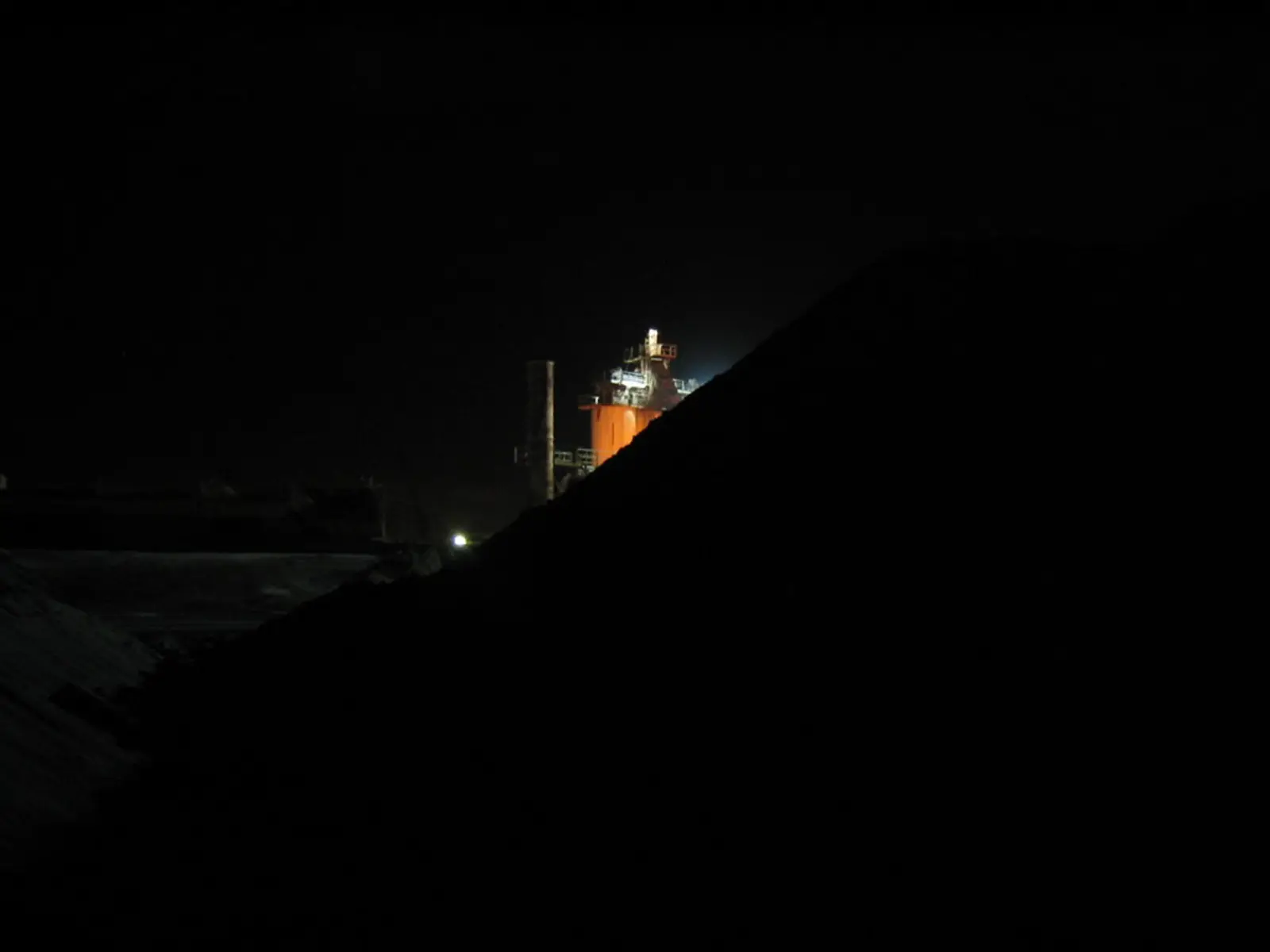 A brightly lit industrial refinery structure is visible at night, partially obscured by a large, dark mound in the foreground.