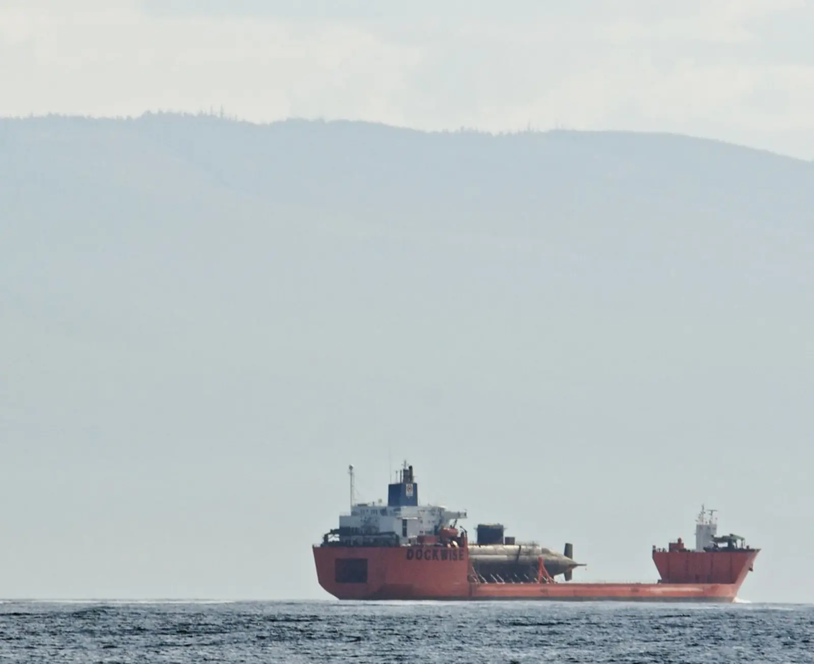 A red heavy-lift ship marked DOCKWISE carries a large, dark, cylindrical object across the water against a backdrop of distant, hazy hills. The photographer titled it “High and Dry”.