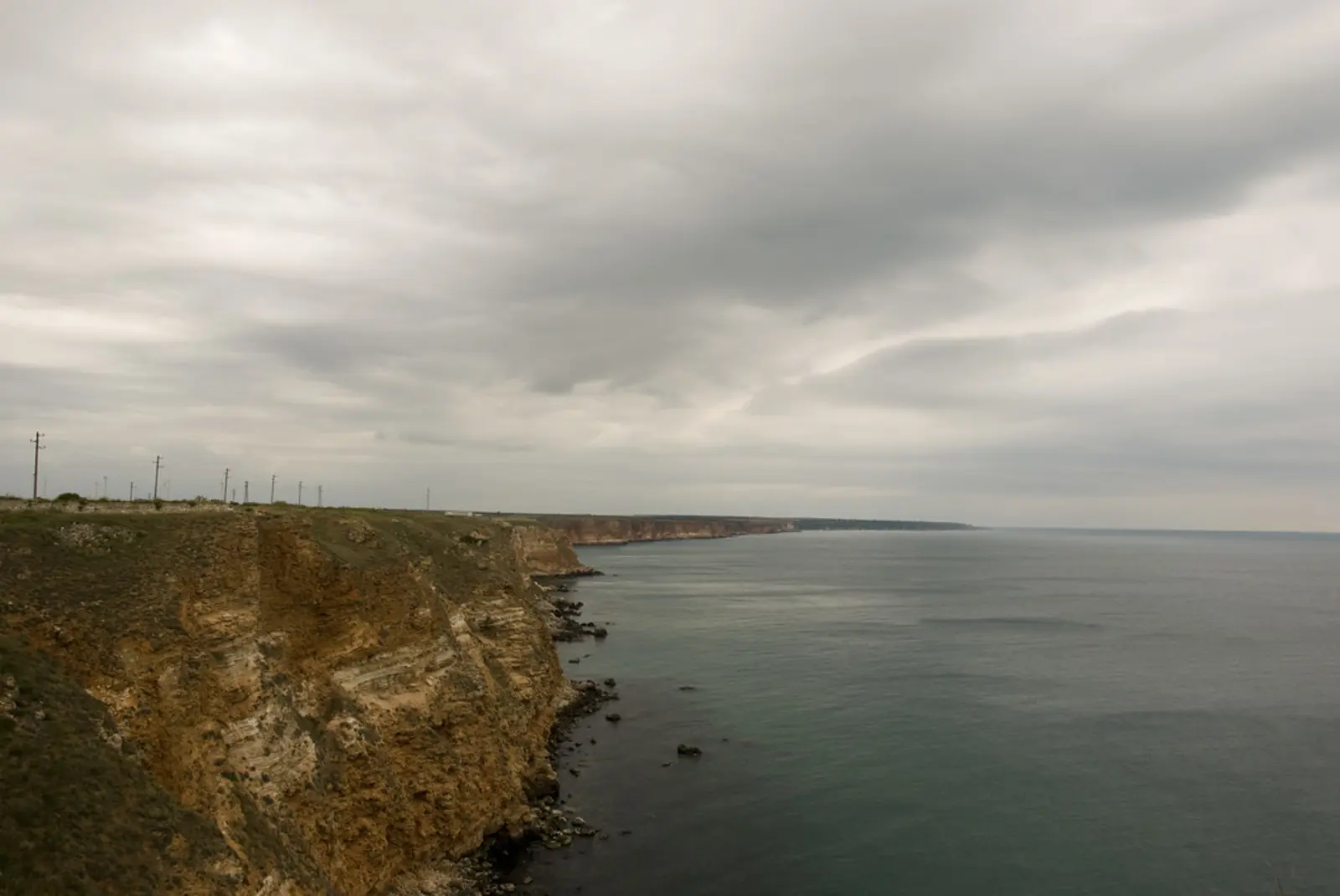 A rugged coastline with steep cliffs extends into the sea under a cloudy, overcast sky, with a line of utility poles visible along the ridge. The photographer titled it “Black Sea coast in Bulgaria”.