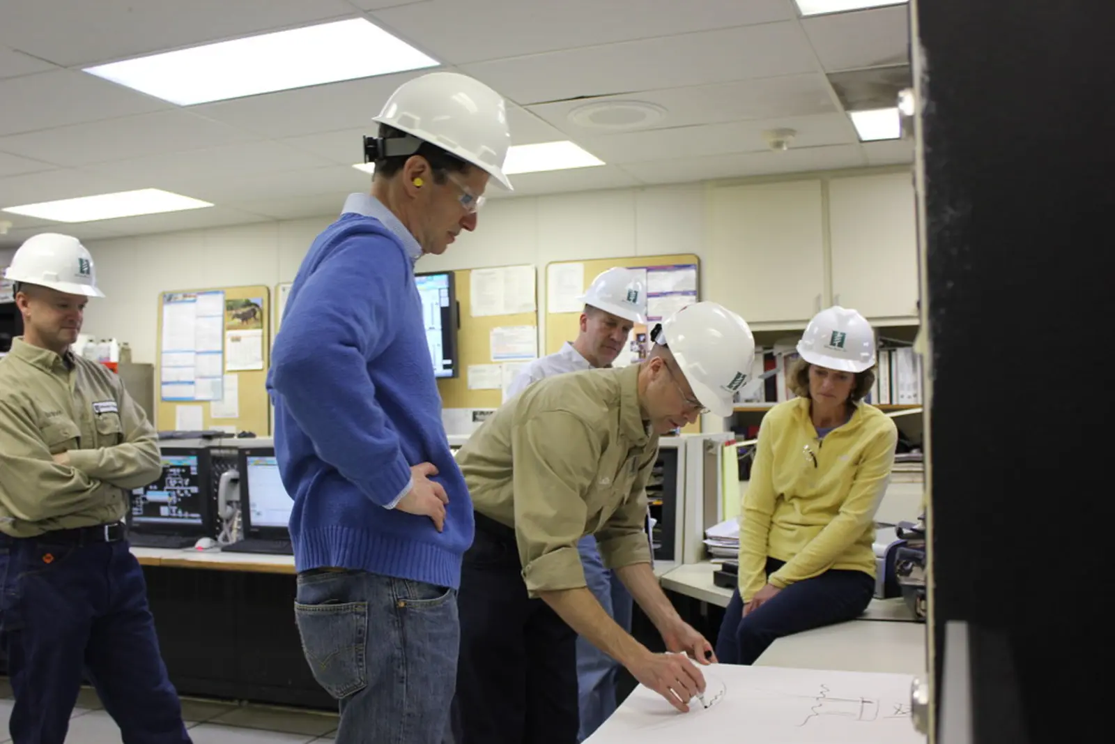 Several people wearing hard hats stand and sit around a table, examining a blueprint in an indoor facility with computer monitors in the background. The photographer titled it “Sen. Wyden and Sen. Murkowski on Cook Inlet offshore oil platform”.