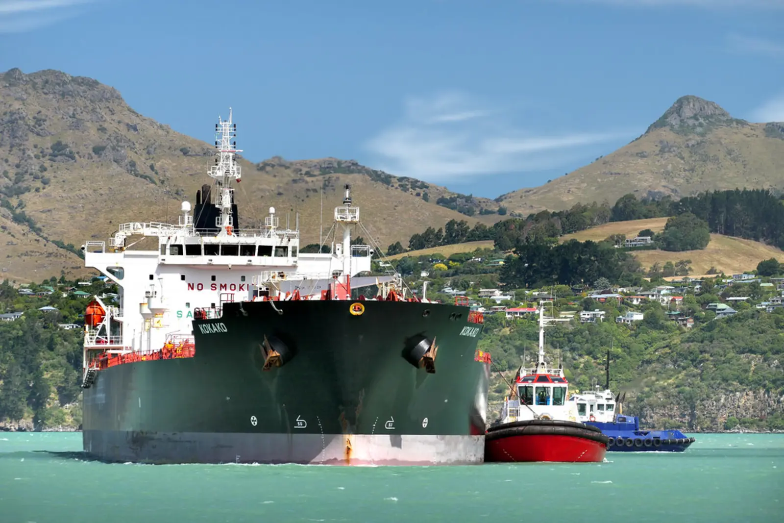 A large dark-hulled tanker ship is moving through greenish water next to a red tugboat, with a hilly coastline in the background. The photographer titled it “KOKAKO Oil Products Tanker.”.