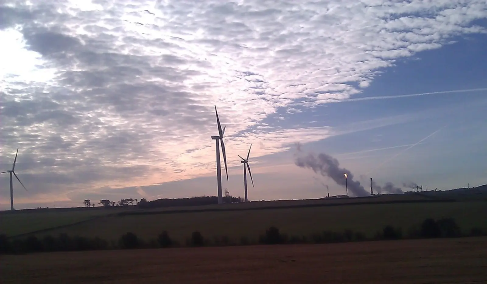 Wind turbines and an industrial facility emitting a plume of smoke or steam stand in a rural landscape under a cloudy sky. The photographer titled it “Wind turbines and oil & gas plant”.